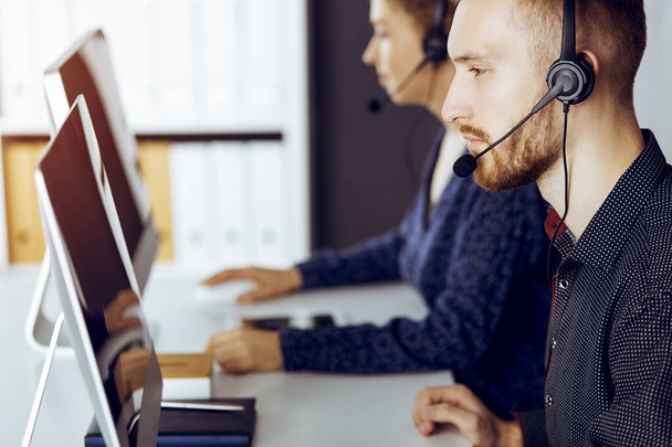 Red-bearded businessman talking by headset near his female colleague while sitting in modern sunny office. Diverse people group in call center - Photo, Image