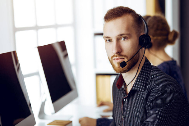 Red-bearded businessman talking by headset near his female colleague while sitting in modern sunny office. Diverse people group in call center - Fotoğraf, Görsel