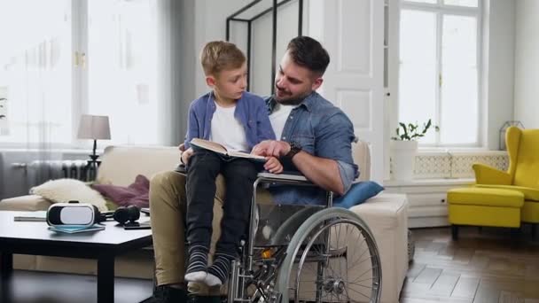 Family concept of good-tempered lucky young bearded disabled dad in wheelchair which helping to read book to his small son that sitting on his knees - Video