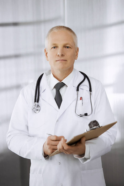 Elderly confident doctor standing straight and using clipboard in clinic. Perfect medical service. Medicine and healthcare concept - Photo, image