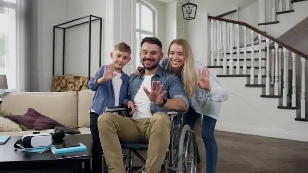 Good-looking loving smiling happy family where young bearded dad sitting in wheelchair looking at camera in modern living-room - Imágenes, Vídeo