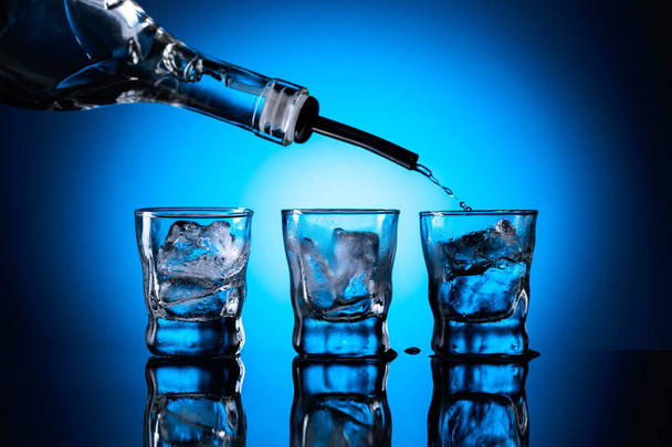 El vodka se vierte de la botella en vasos con hielo. Gafas congeladas sobre fondo azul oscuro. Copiar espacio. - Foto, Imagen