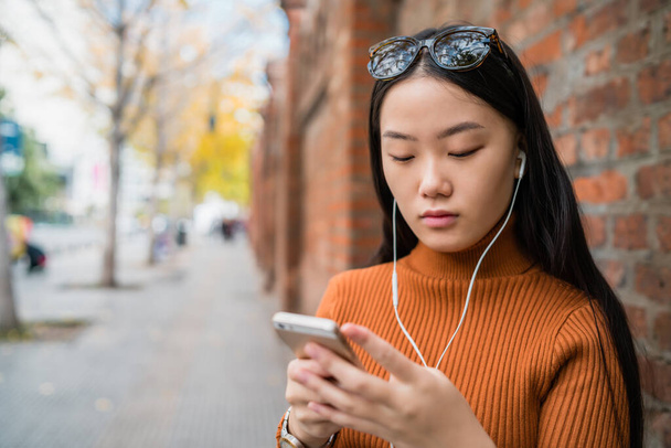Portrait of young Asian woman using her mobile phone with earphones outdoors in the street. Urban and communication concept. - Фото, изображение