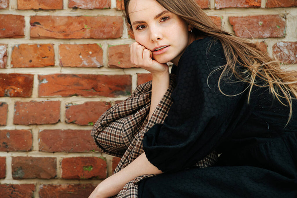 Side portrait of a crouching young woman in a long black dress in front of a old house brick wall. Holding checkered jacket. Cropped at the top. - Photo, Image