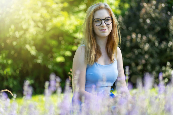 Smiling young woman is posing with glasses behind lavender in the garden - Фото, изображение