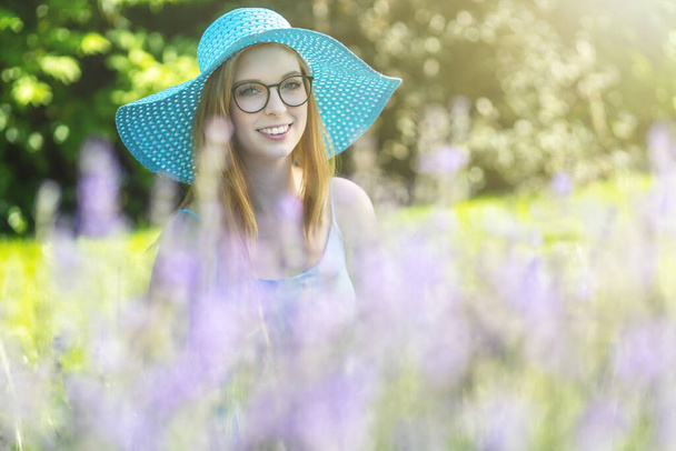 Portrait of smiling young woman with glasses wearing turquoise summer hat is posing behind lavender in the garden - Foto, immagini
