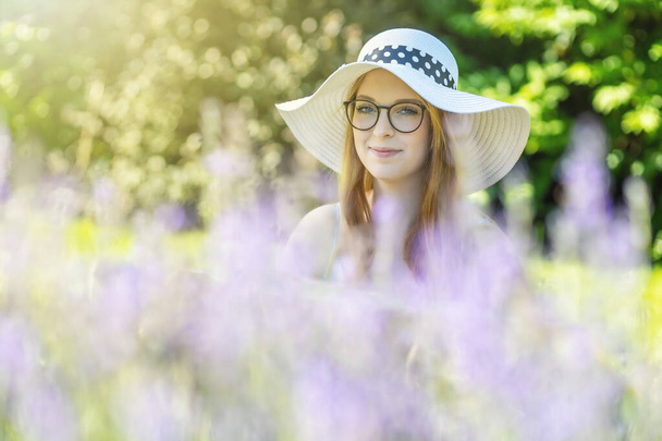 Portrait of smiling young woman with glasses wearing white  summer hat is posing behind lavender in the garden - 写真・画像