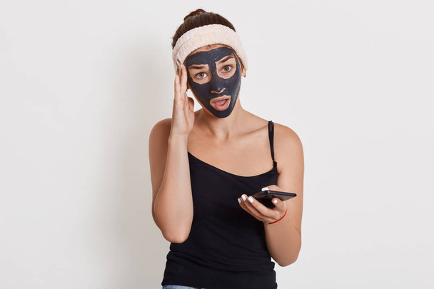 Charming young lady with black facial cosmetic mask using cellphone, looking directly at camera, keeping finger near temple, having astonished and puzzled facial expression. - Foto, Imagem