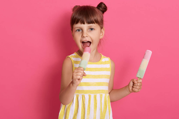 Little girl holding and eating two ice cream, keeping mouth widely opened and biting water ice, looking directly at camera, wearing dress with white and yellow stripes. - Valokuva, kuva