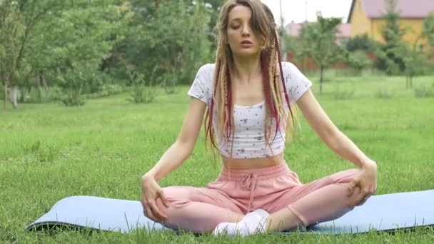 Joven hermosa mujer con rastas está haciendo yoga sentado en posición de loto al aire libre durante la mañana en la hierba verde de su patio trasero - Imágenes, Vídeo