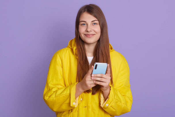 Tevreden mooie vrouw houdt moderne mobiele telefoon, sms 'jes met vriend online in de chat, geklede stijlvolle gele shirt, kijken direct naar de camera, staat tegen lila muur. - Foto, afbeelding