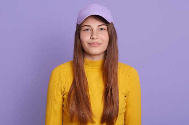 Close up portrait of tender charming girl, posing smiles, standing in baseball hat isolated over lilac background, wearing yellow casual shirt, being in good mood. - 写真・画像