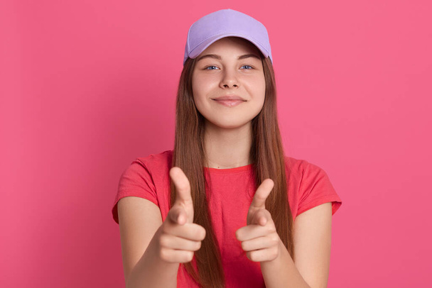 Cheerful lovely girl pointing to camera with pleased facial expression, has satisfied look, wearing red t shirt and baseball cap, posing isolated over pink background. - Foto, Bild