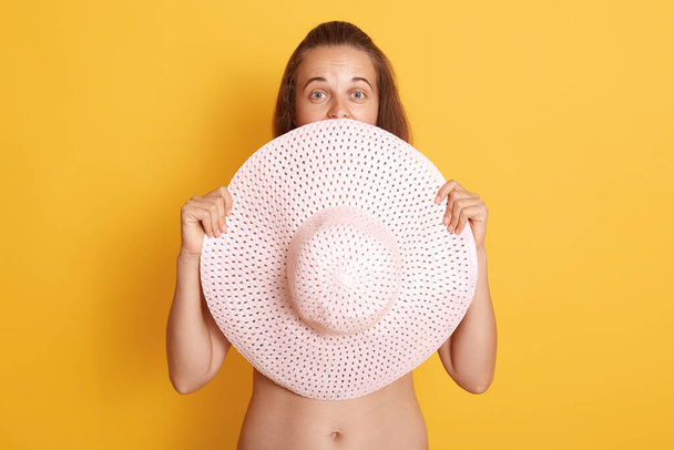 Attractive young brunette woman without clothes hide her boobs behind straw hat, sowing her bare belly, looking at camera isolated over yellow background. - Fotografie, Obrázek