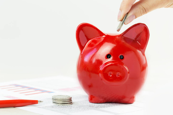 Fingers putting coin into red piggy coins bank with financial papers on desk. Savings, insurance. Business or home finance. Credit or income concept with copy space - Fotó, kép