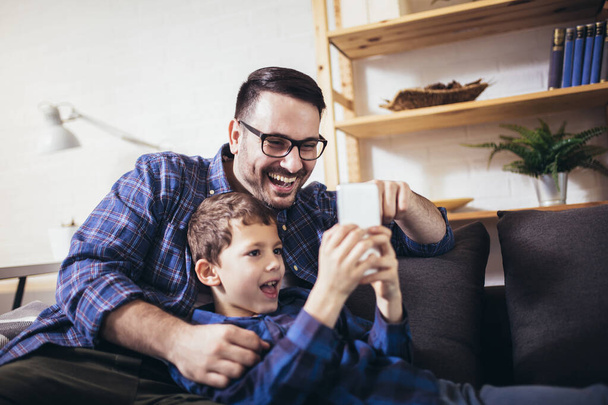 Fiatal apa öleli kisfia család ül a kanapén otthon segítségével okos telefon úgy érzi, boldog vidám nevetés vicces videók. - Fotó, kép