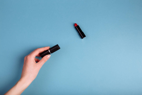 Lady's hand holding closed red lipstick isolated on blue background near to opened lipstick. - Photo, Image