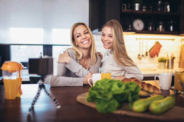 幸せな母親と娘とともにスマートフォンを持って朝食を取り、自宅のキッチンで自撮り - 写真・画像