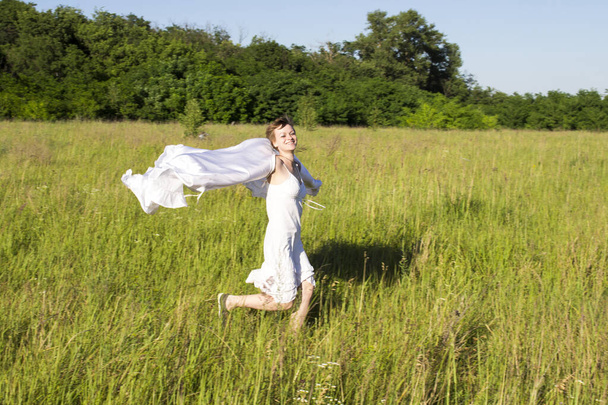 Une fille dans une robe blanche, court à travers le champ, avec un chiffon blanc dans ses mains et rit - Photo, image