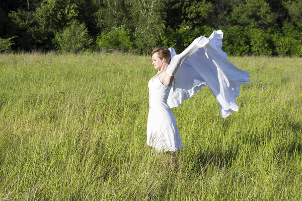 Une fille en robe blanche se tient debout sur le terrain et tient un tissu blanc flottant dans ses mains - Photo, image