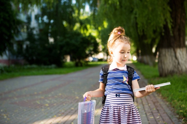 A little girl of Caucasian appearance in a school uniform with a backpack and the book and a set of markers. back to school. Elementary school, developing activities for preschoolers. Space for text - Φωτογραφία, εικόνα