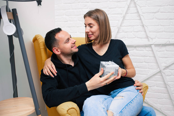 Portrait of couple young in a cozy room. A man sits on a yellow chair. A woman sits on the lap of a man and holds a gift Christmas family holidays. Modern style interior.  - Zdjęcie, obraz