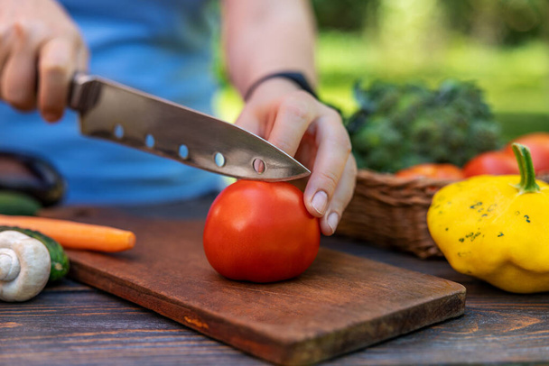 Una mujer está cortando verduras, las manos de una mujer están cortando un tomate en una ensalada en una mesa de madera, varias otras verduras se encuentran cerca. Concepto de alimentos orgánicos saludables. - Foto, Imagen