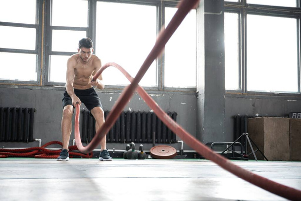 Athletic young man with battle rope doing exercise in functional training fitness gym. - Foto, Imagen