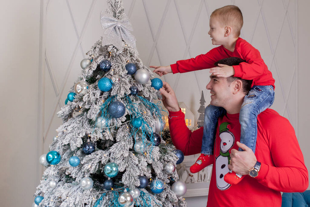 Happy Father and son enjoying decorating Christmas tree with Christmas balls and light garland preparing for celebrating winter holidays: Merry Christmas Eve and Happy New Year 2021. Family concept. - Fotó, kép