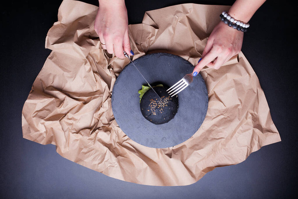 Zwarte broodrol met zwarte sesam in handen gehouden over een kraftpapier in menselijke handen zwarte achtergrond. Boven het uitzicht van vreemde zwarte hamburger brood. Halloween eten - Foto, afbeelding