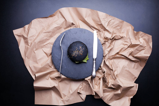 Μαύρο ρολό ψωμιού με μαύρο σουσάμι που κρατιέται στα χέρια πάνω από ένα χαρτί κραφτ σε ανθρώπινα χέρια μαύρο φόντο. Πάνω από την άποψη των παράξενο μαύρο ψωμί χάμπουργκερ. Απόκριες τροφίμων - Φωτογραφία, εικόνα