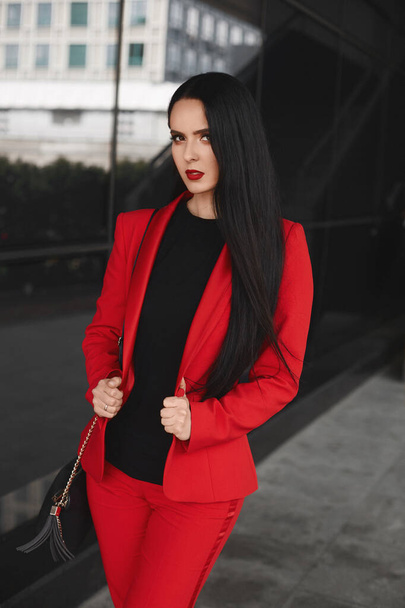 Gorgeous brunette woman with black hair in an elegant red suit looking at the camera and posing outdoors - Φωτογραφία, εικόνα