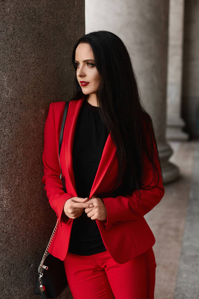 Young businesswoman with red lips and in the red stylish suit posing outdoors on the city street - Photo, Image