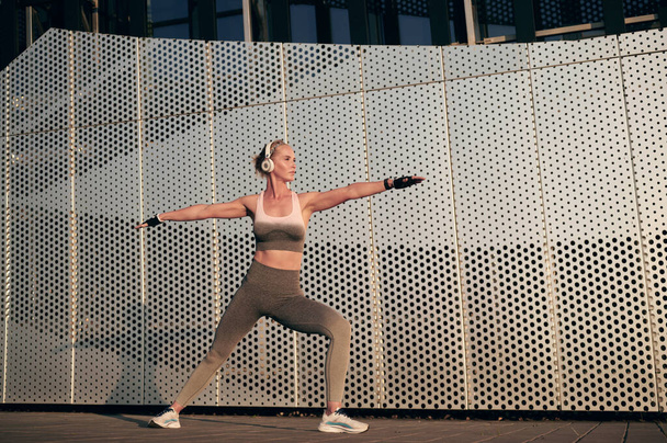 Ajuste fuerte hembra en auriculares y ropa de fitness está haciendo ejercicio de estiramiento después del entrenamiento al aire libre en frente de la pared - Foto, Imagen