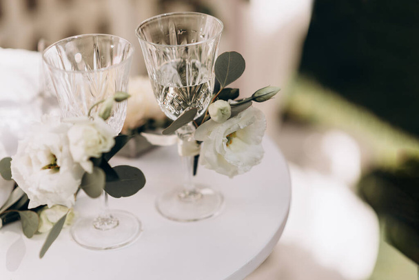 Twee glazen kristal voor een bruidspaar. bril versierd met witte bloemen staan op een witte tafel - Foto, afbeelding