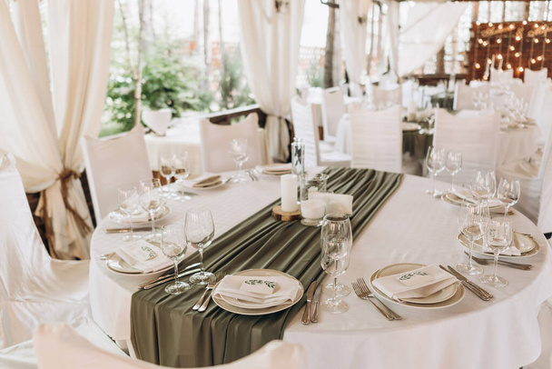 Wedding table setting. Banquet table with white tablecloths served with white plates, silver cutlery, white napkins and wine glasses. olive colore drapery on a white set table - Foto, imagen