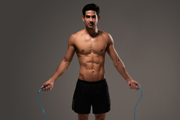 Attractive muscular young man doing workout with jump rope against gray background - Foto, Imagen