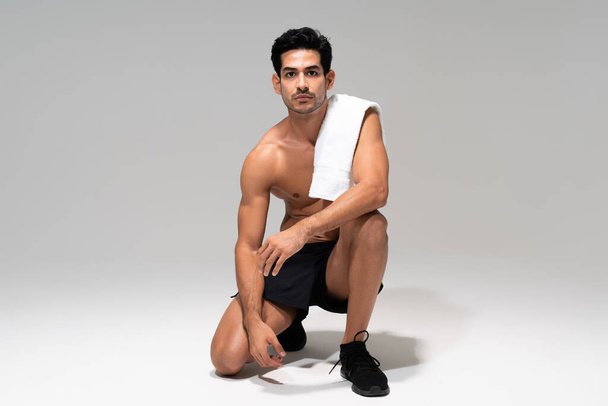 Jeune homme latin athlétique avec serviette sur l'épaule à genoux dans un studio avec fond gris - Photo, image