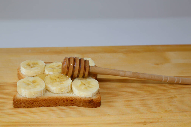 Auf einer Scheibe Brot befinden sich Bananenstücke mit Honig auf dem Hintergrund der Holzoberfläche. Auf einem Stück Brot mit aufgeschnittenen Früchten und mit med. Das Sandwich liegt auf dem Holztisch. - Foto, Bild