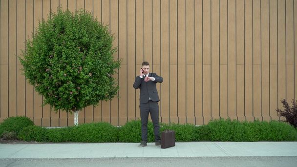 Молодий бізнесмен у костюмі говорить по телефону. Серйозний чоловік стоїть з портфелем в руці на фоні будівлі з клумбою
. - Фото, зображення