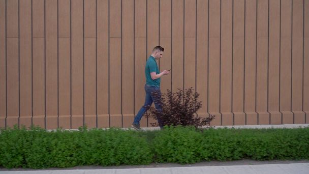 Ένας νεαρός άνδρας πηγαίνει με ένα τηλέφωνο στο χέρι του. Ένας άντρας περπατάει κοντά στο κτίριο και συνεχίζει την αλληλογραφία του σε ένα smartphone.. - Φωτογραφία, εικόνα