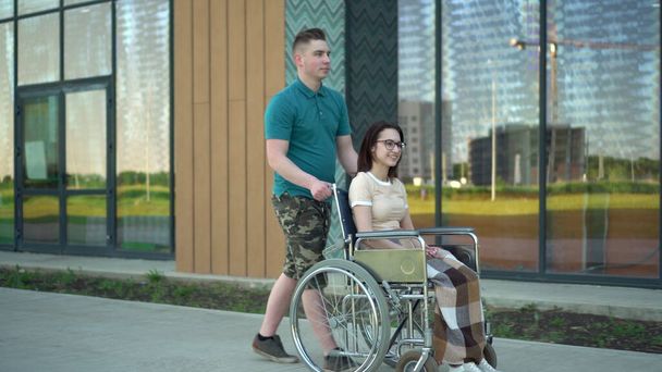 Egy fiatalember egy fiatal nőt cipel tolószékben. Egy asszisztens tolószékben cipel egy mozgássérültet az utcán. Speciális szállítás a fogyatékkal élők számára. - Fotó, kép