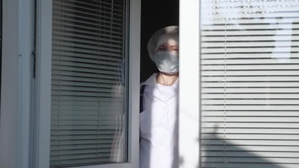 Una doctora cansada con traje protector mira por la ventana, se quita la máscara médica, cierra los ojos por fatiga, respira aire fresco. Día de trabajo duro de médico en el hospital. Coronavirus pandémico - Imágenes, Vídeo