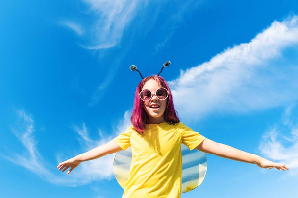 Girl in a bee costume with pink hair outdoors. Looks into the camera Child pretends fly. Blue sky background Copy space - Foto, Imagem