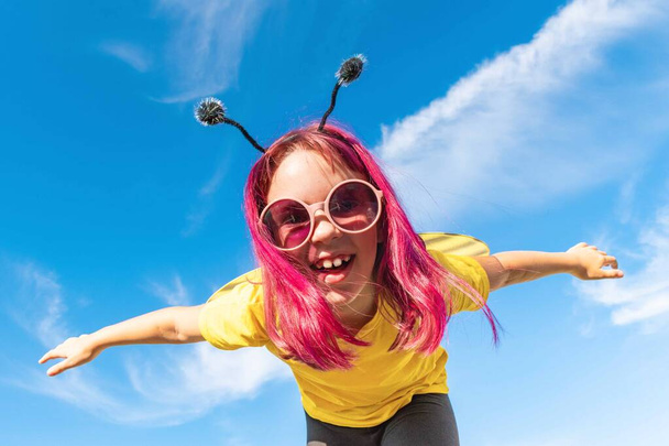 Girl in a bee costume with pink hair outdoors. Looks into the camera Child pretends fly. Blue sky background Copy space - Foto, immagini