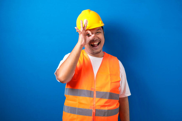 Agradable seguro de grasa asiática constructuin trabajador hombre con amplia sonrisa, con chaleco naranja y casco de seguridad muestra gesto bien contra fondo azul - Foto, imagen