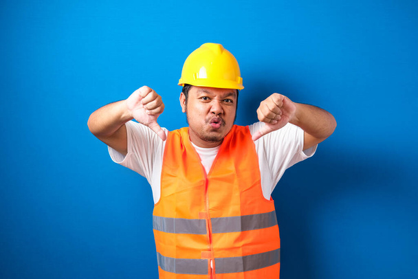 オレンジ色の安全ベストとヘルメットを着て若い脂肪アジアの建設労働者の男性は、青の背景に対して親指ダウンジェスチャーで不満と否定を示し、怒っているように見えます。悪い表現. - 写真・画像