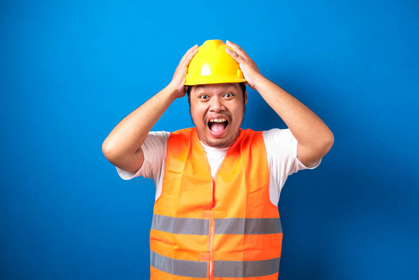 zszokowany tłuszcz azjatycki budowniczy pracownik człowiek jest trzymając jego ręce na jego głowie z szerokim otwartymi ustami odizolowane na niebieskim tle. - Zdjęcie, obraz