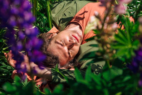 Tall handsome man in a green jacket lying among the lupines flowers. Top view. - Foto, imagen