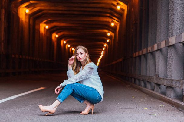 Young woman with long blond hair sitting next to the dark industrial tunnel with lights - Photo, image